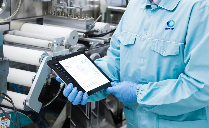 Operator in a cleanroom coat and gloves reviewing a safety dashboard on a tablet beside a packaging production line